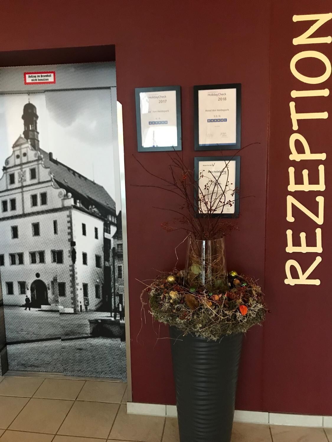 Un espace d’accueil agrémenté d’une photographie encadrée d’un bâtiment, de deux certificats encadrés et d’une grande plante en pot.