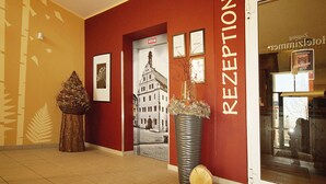 Zona de recepción con pared roja, cuadros enmarcados, un jarrón grande con flores y una puerta que da a una habitación.