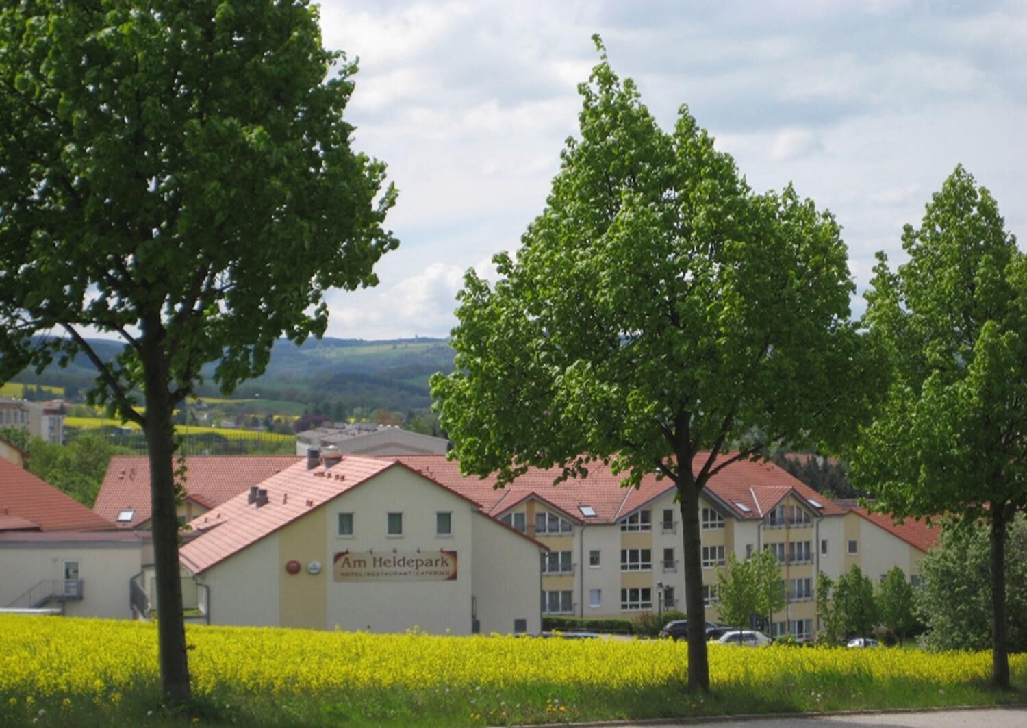 Um hotel com uma placa que diz "Am Heidepark Hotel Restaurant Catering". Possui telhado de telhas vermelhas, várias janelas e é cercado por árvores verdes e um campo de flores amarelas.