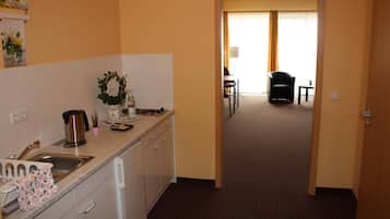 Et hotelkøkken med vask, komfur og skabe. Et spiseområde med et bord og stole kan ses igennem en åben dør.