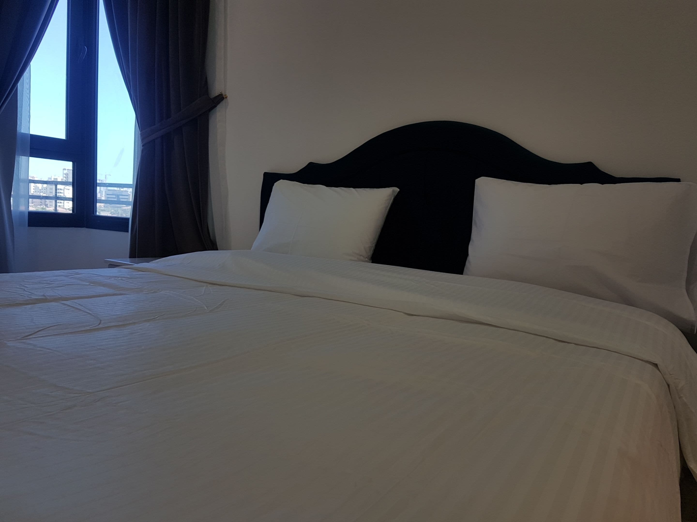 Una cama con ropa de cama blanca y un cabecero oscuro en una habitación con ventana y cortinas.