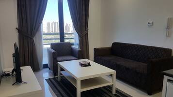 Una sala de estar moderna con una mesa de centro blanca, un sofá oscuro con estampado y un sillón gris. Hay una ventana con cortinas, una pared blanca y una pequeña mesa blanca con una computadora y una regleta.