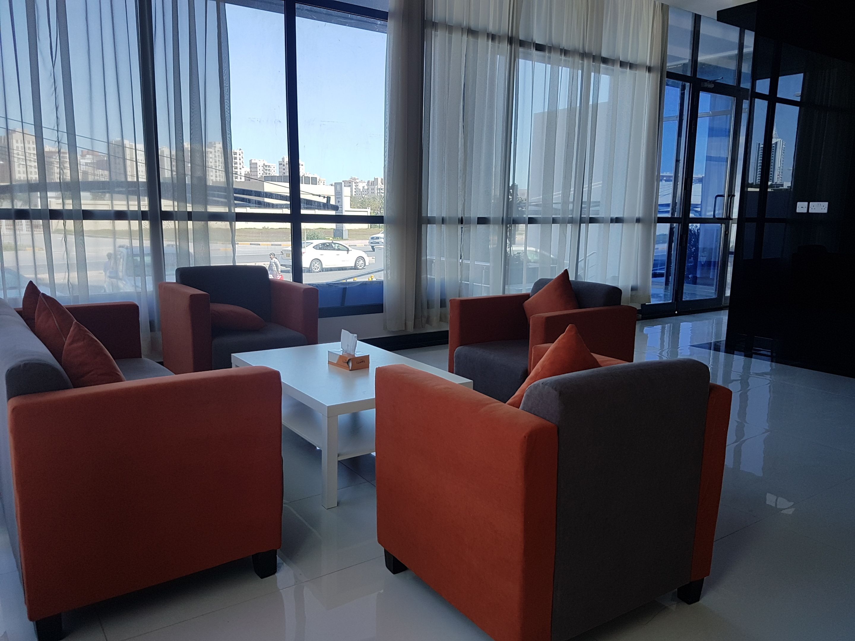 A modern hotel lobby with large windows, orange and grey sofas, a white coffee table, and a view of the cityscape.