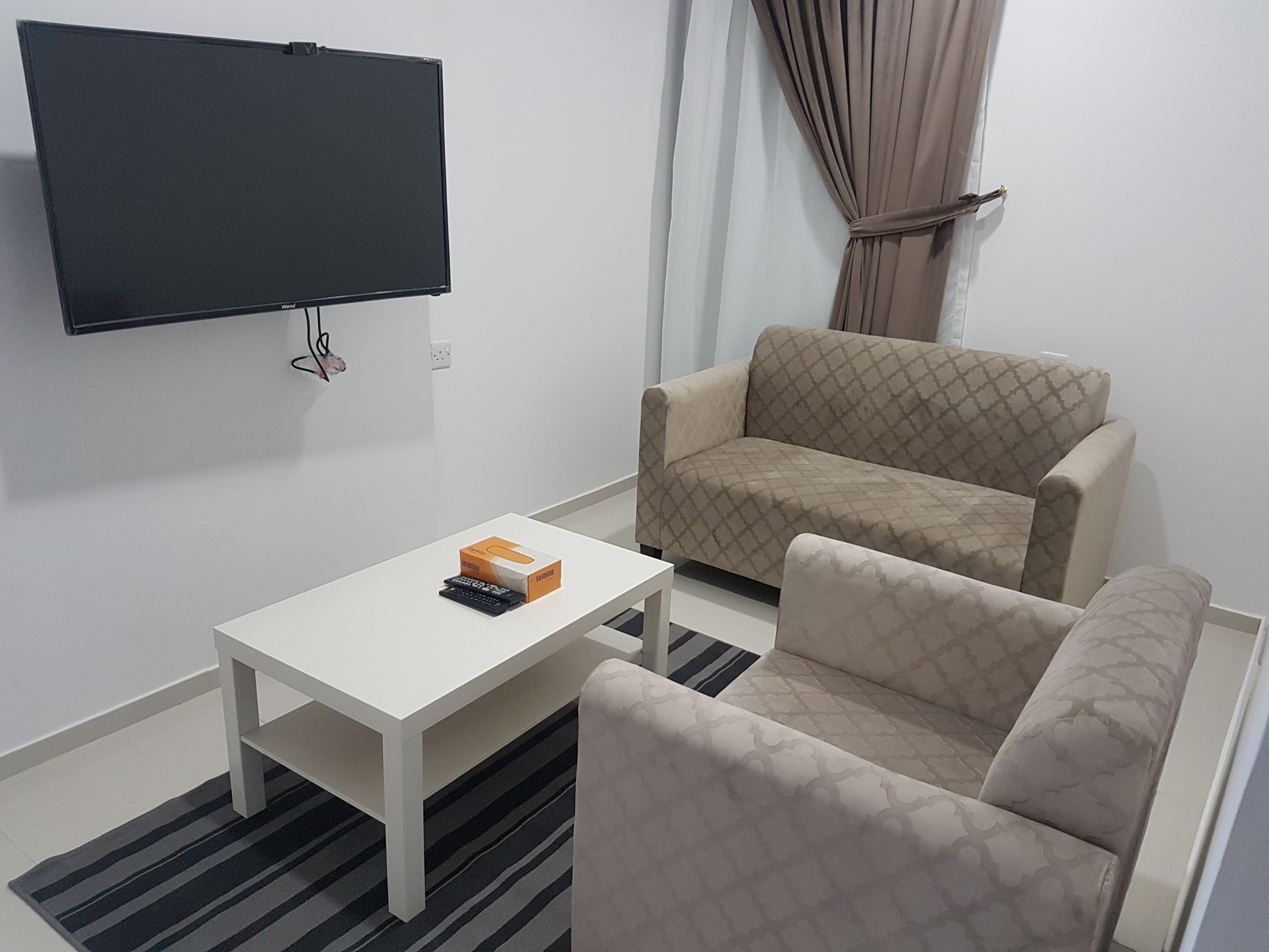 Un salon avec un téléviseur à écran plat fixé au mur, une table basse blanche, un canapé beige à motifs et un tapis rayé.