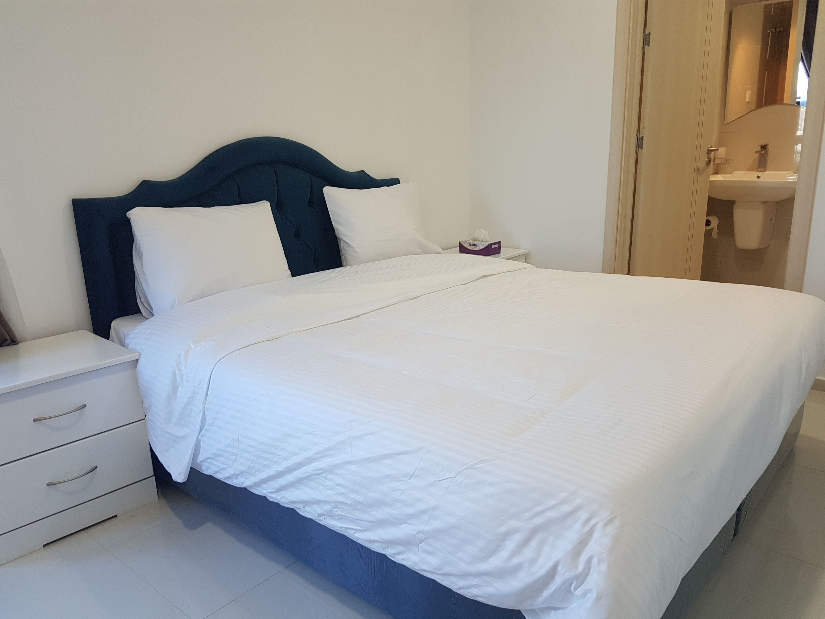 A neatly made bed with white bedding and a dark headboard, a white bedside table with drawers, and a visible bathroom with a sink and mirror.