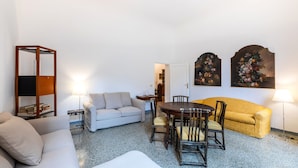 Ein Wohnzimmer mit einer Couch, einem Esstisch mit Stühlen, einem Bücherregal und zwei Wandbildern.