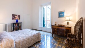 Ein Schlafzimmer mit Bett, Schminktisch, Stuhl, Heizkörper, Bücherregal und einer Tür, die zu einem Außenbereich führt.