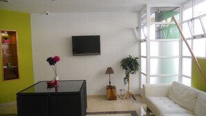 Een woonkamer met een zwarte salontafel, een witte bank, een aan de muur bevestigde televisie en een raam met jaloezieën.
