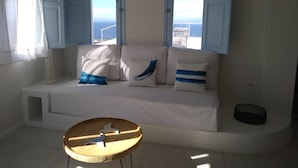 A room with a white sofa, two windows with blue shutters, a small round wooden table, and a view of the sea. - Sol Mate Suites (Santorini)