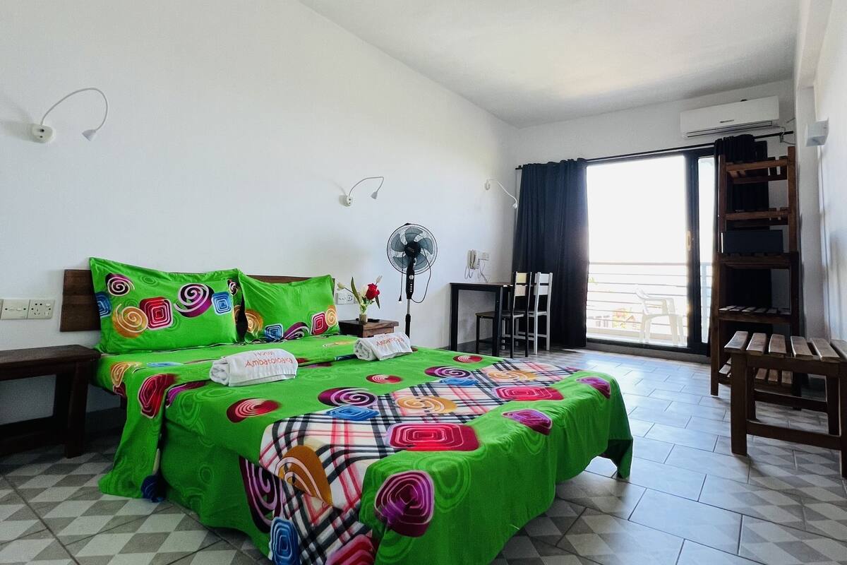 Habitación de hotel con piso a cuadros, cama con ropa de cama verde, escritorio, silla, ventilador y vista al exterior.