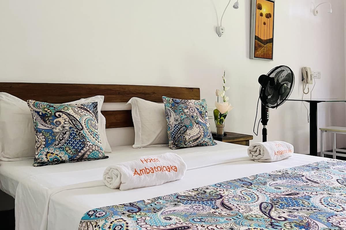 Habitación de hotel con cama, dos almohadas estampadas, una mesita de noche con un ventilador, un jarrón con flores y un cuadro colgado en la pared.