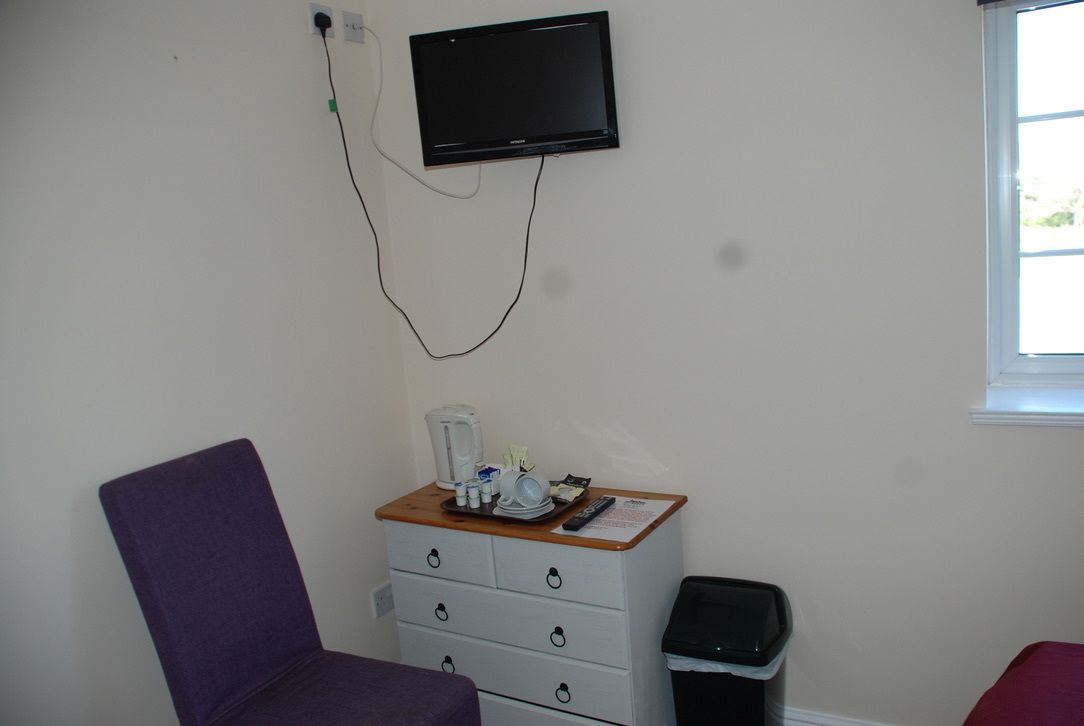 Una habitación con una televisión de pantalla plana montada en la pared, un aparador de madera con artículos de aseo, una silla morada y un cubo de basura.