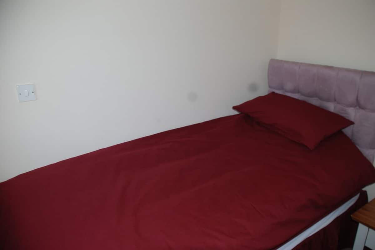 A single bed with a red bedspread and a matching pillow, a white wall socket, and a wooden nightstand.