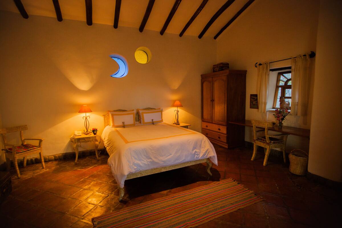 Een slaapkamer met een groot bed, twee nachtkastjes met lampen, een houten kledingkast, een stoel en een raam met gordijnen.