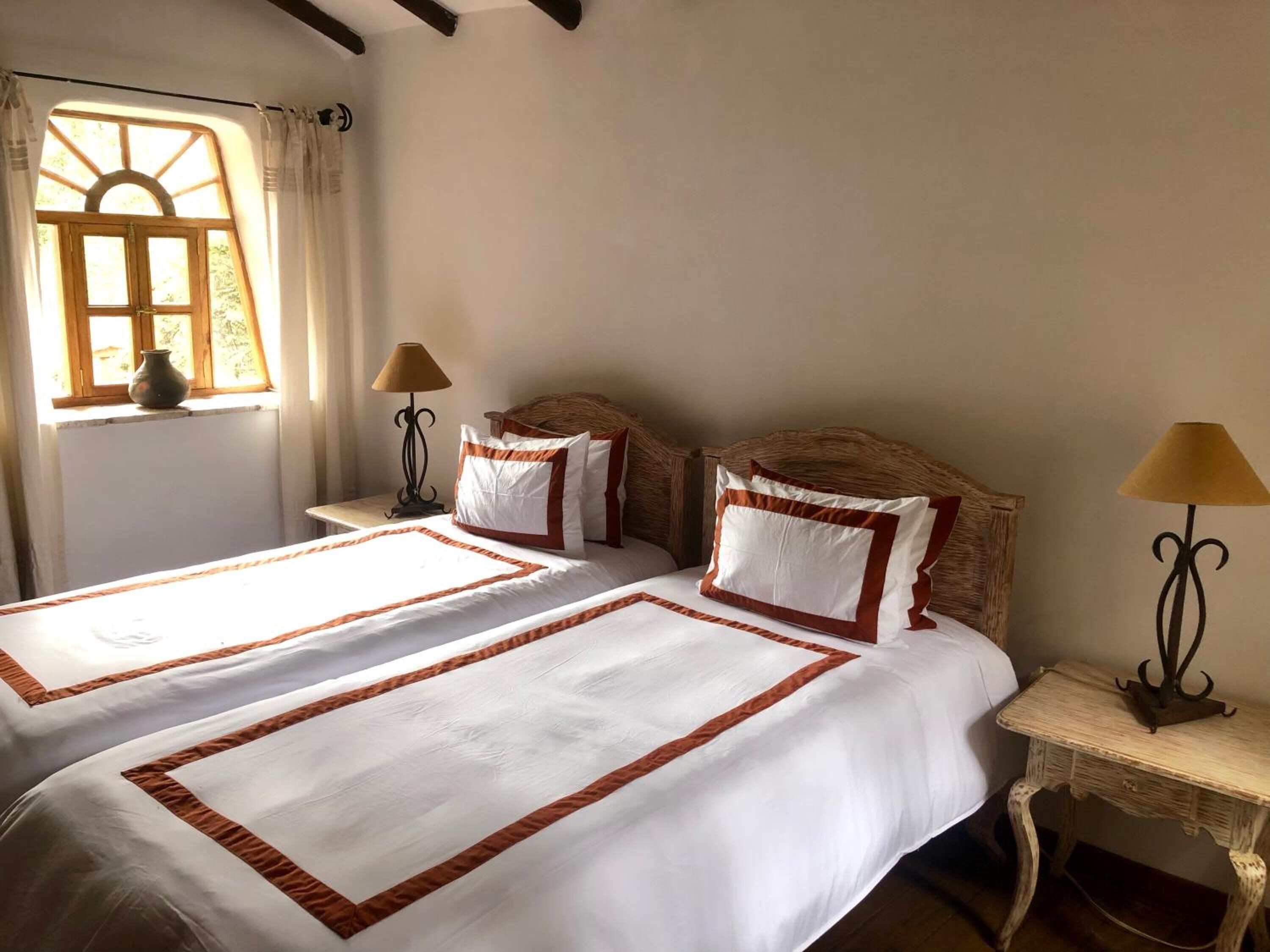 Cama doble con ropa de cama blanca y detalles marrones, ventana con cortinas y una lámpara sobre una mesita de noche.