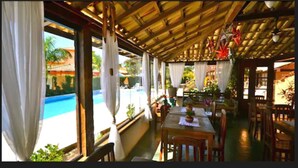 A covered outdoor dining area with a pool view, wooden furniture, and a thatched roof. - Da Ferradura (Búzios)