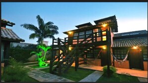 A traditional wooden house with a staircase leading to a second floor, surrounded by greenery and a hammock. - Da Ferradura (Búzios)