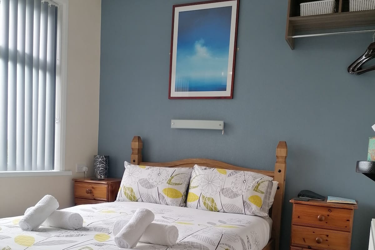 Une chambre à coucher bien rangée, avec un lit, une commode en bois, une table de chevet et une étagère ornée d’objets décoratifs.