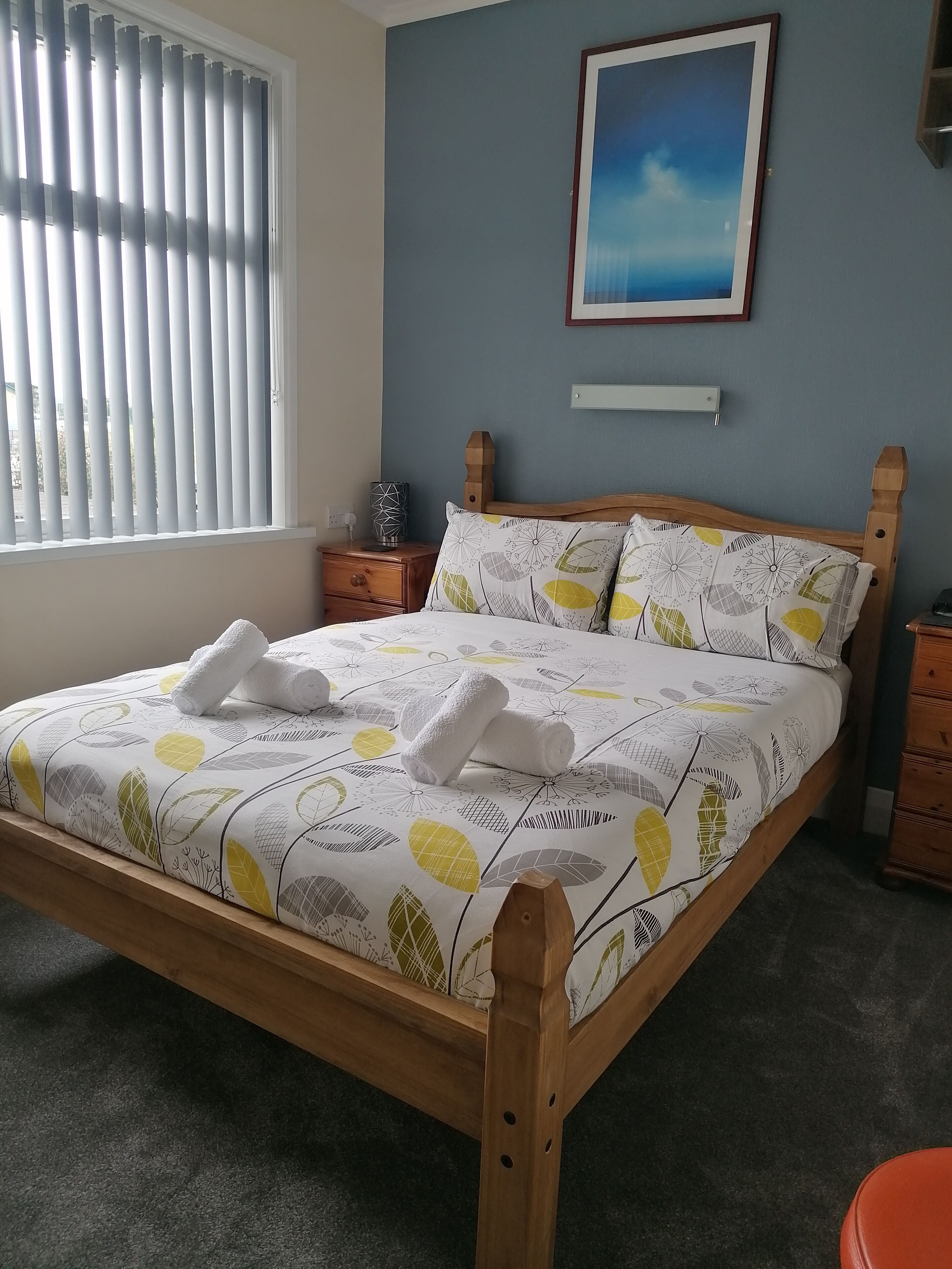 Un lit bien fait, recouvert d’une couvre-lit à motifs de feuilles, avec une tête de lit en bois et deux oreillers.