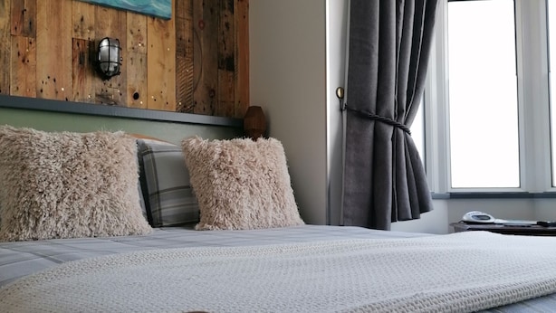 A bedroom with a wooden headboard, a large bed with a white and blue plaid duvet, fluffy pillows, and a painting of a coastal scene on the wall.