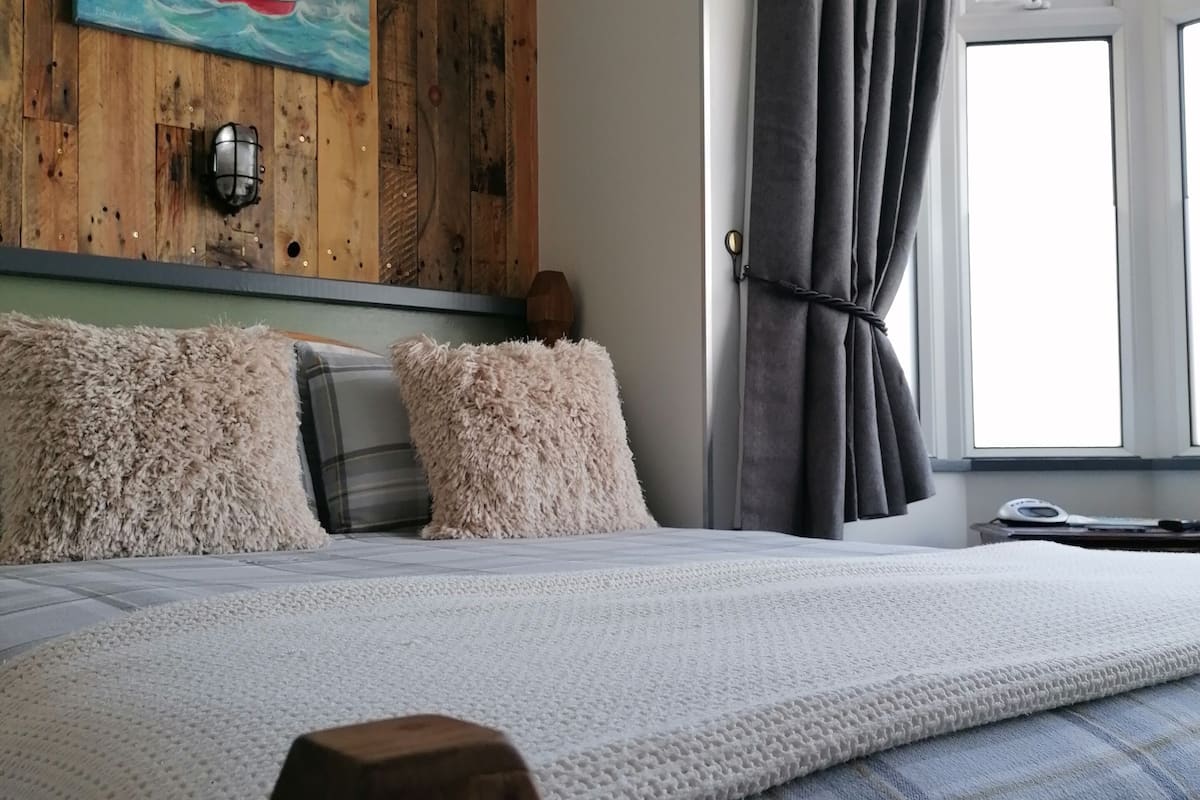 Une chambre à coucher avec une tête de lit en bois, un grand lit recouvert d’une couette à carreaux blancs et bleus, de moelleux oreillers et un tableau représentant un paysage côtier accroché au mur.