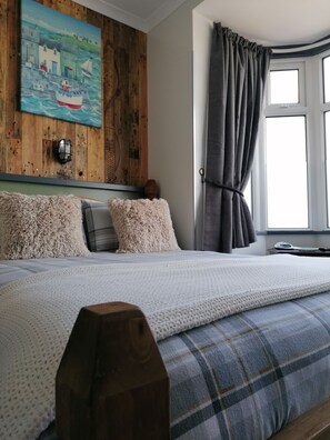 Un dormitorio con cabecera de madera, una cama grande con edredón azul y blanco escocés, almohadas mullidas y un cuadro de una escena costera en la pared.