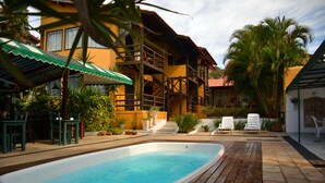 Una piscina con plataforma de madera, un área de descanso techada y un edificio con balcones y macetas con plantas.
