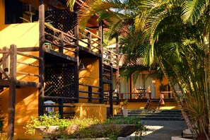 A wooden balcony with a lattice design, a yellow building, and a palm tree. - Kilandukilu (Búzios)