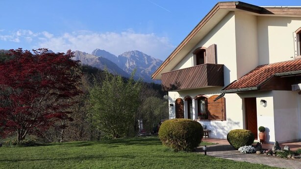 A house with a well-maintained lawn, a wooden balcony, and a mountain backdrop. - Bed & Breakfast La Pecora Nera (Belluno)