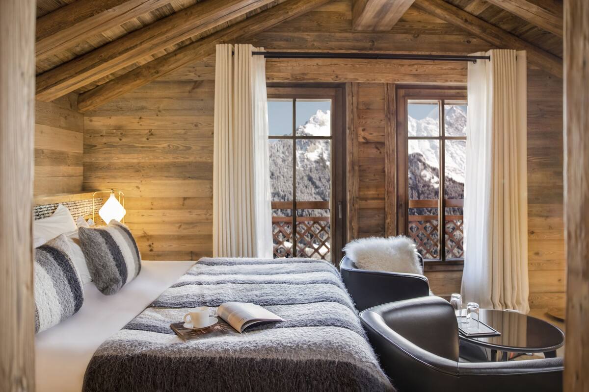 Acogedora habitación tipo cabaña de madera con cama, una silla, una mesita y ventana con vistas a la montaña.