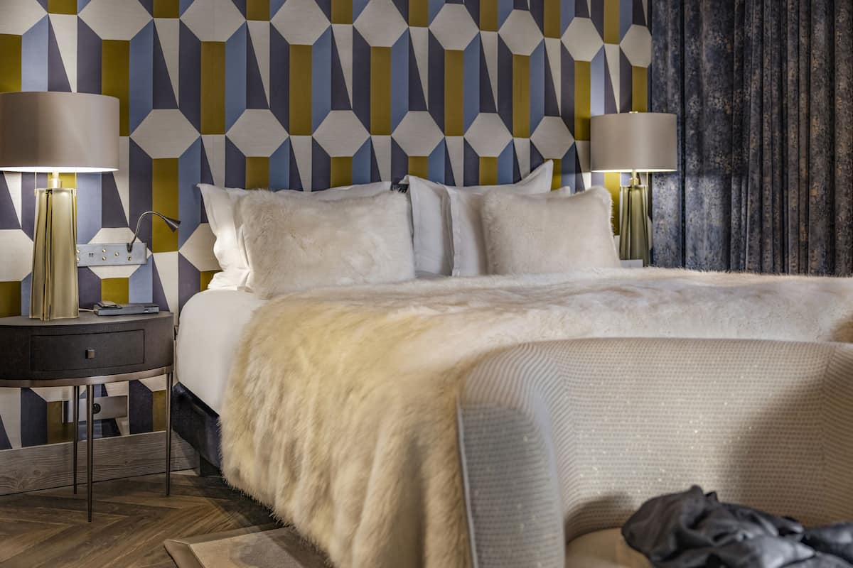 Una habitación de hotel moderna con pared de diseño geométrico, una cama grande con ropa de cama blanca, dos lámparas de noche y una mesita de noche con lámpara.