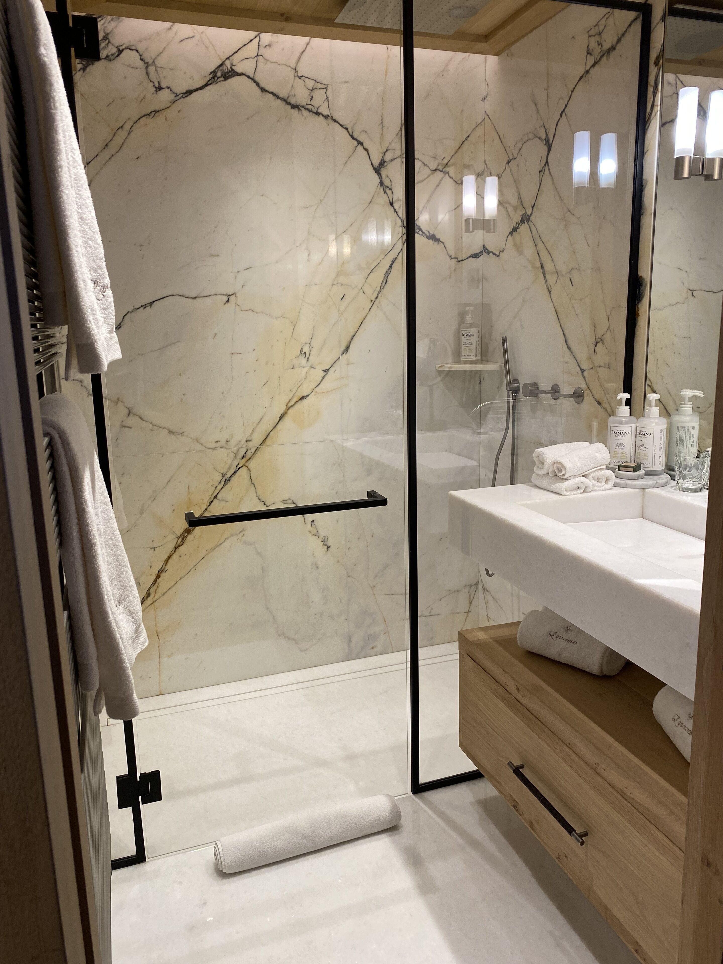 A modern bathroom with a glass-enclosed shower, marble walls, a wooden vanity, and neatly arranged toiletries.