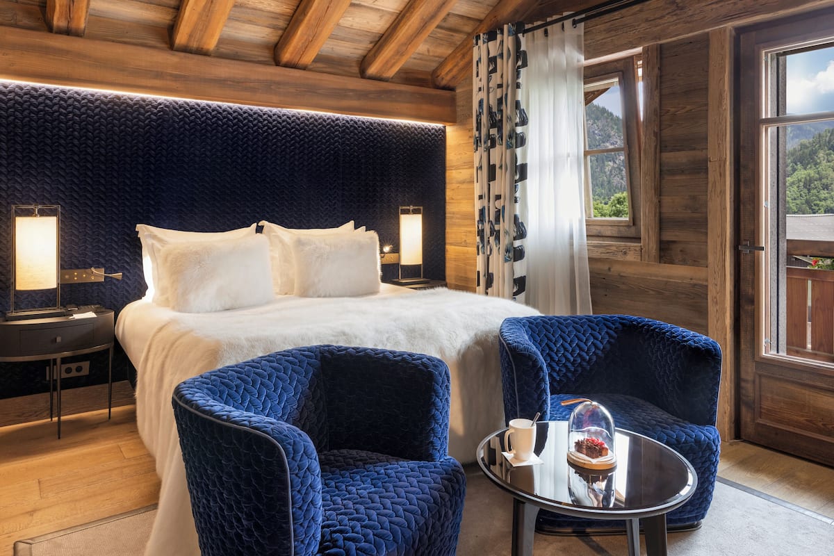 Una habitación acogedora con una cama grande, dos sillones azules, una mesita con una taza y un vaso, y una ventana con cortinas.