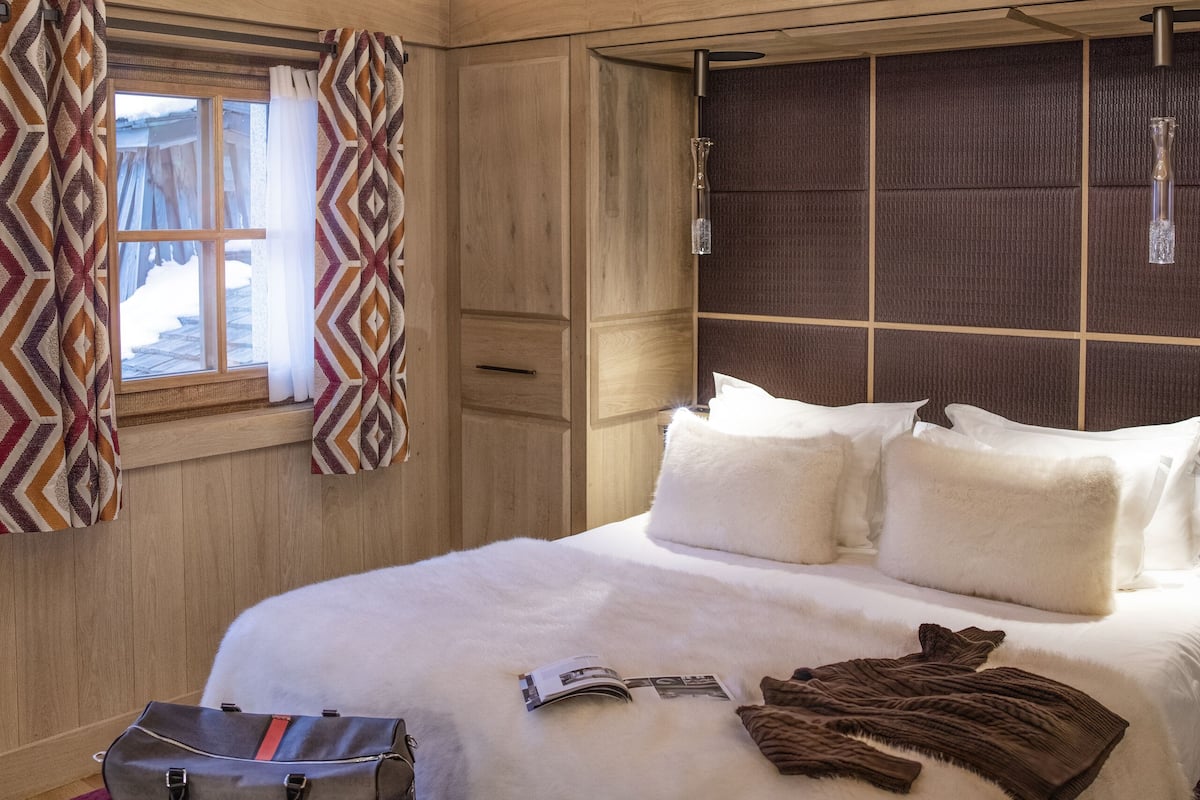 Un dormitorio con techo de madera, una araña, una cama con ropa de cama blanca, un banco y una ventana con cortinas estampadas.