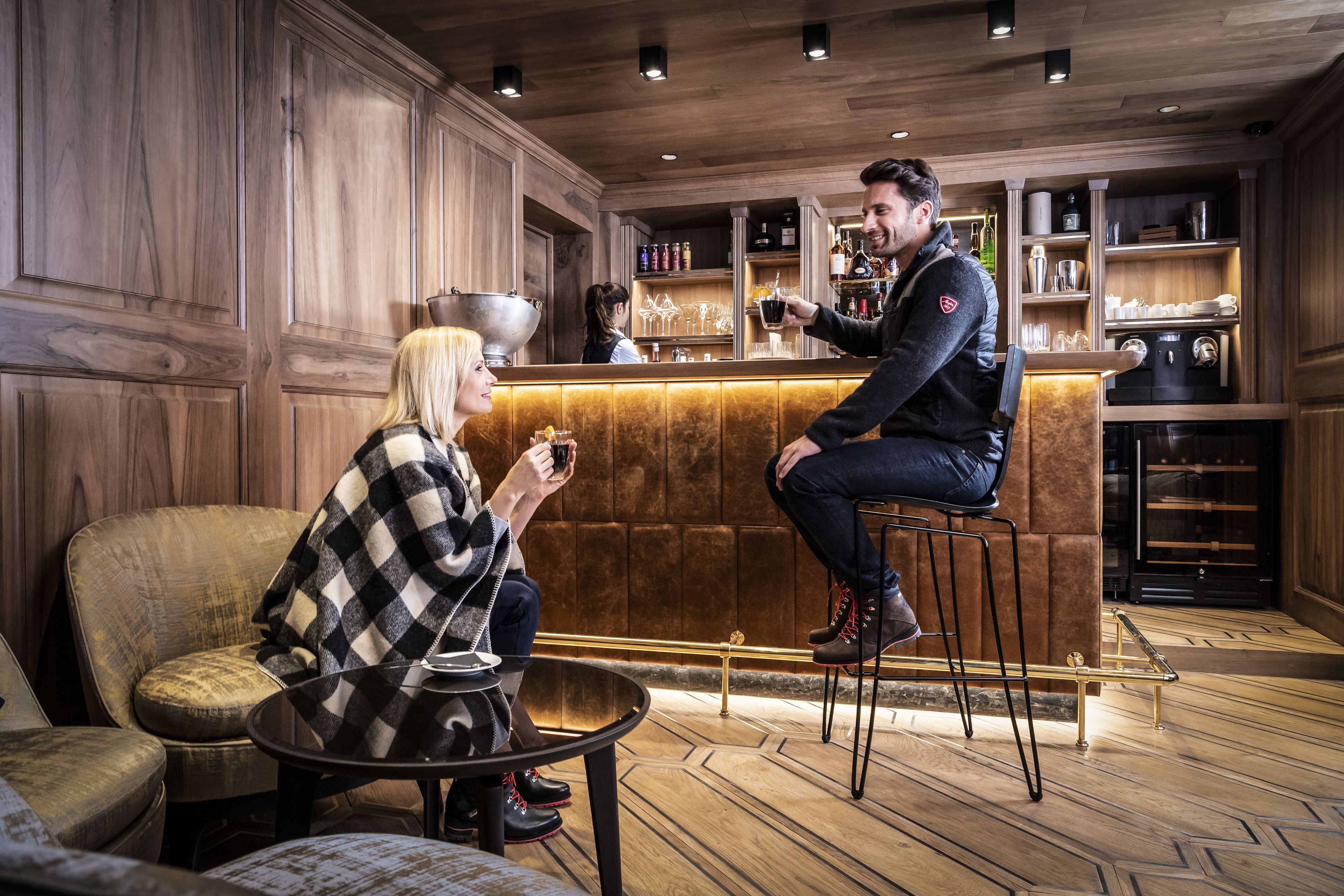 Een bar met houten lambrisering, een toog en planken met flessen en glazen.