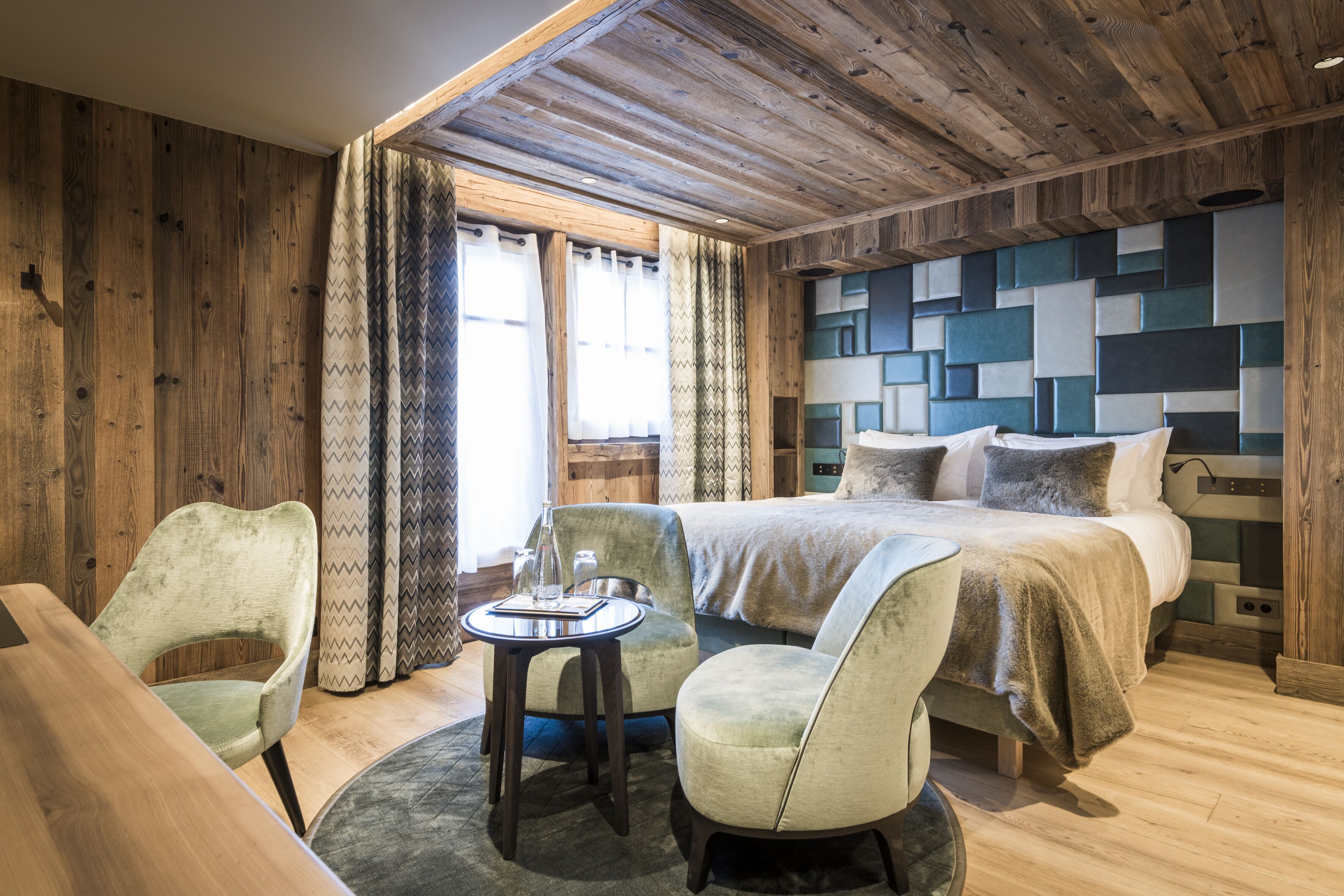 A cozy room with a wooden ceiling, a bed with a geometric headboard, two upholstered chairs, and a small round table.