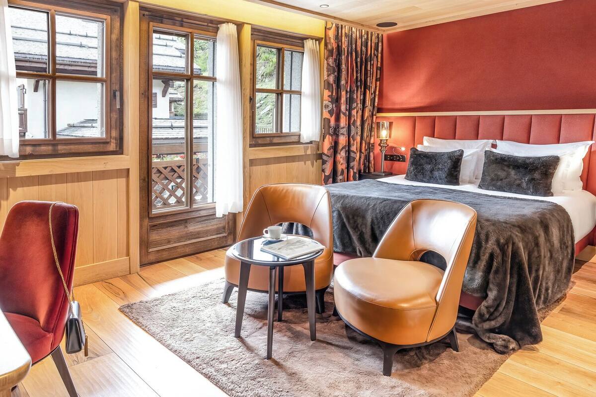 Habitación de hotel con una cama grande, dos sillones de cuero, una mesita y vistas a un edificio tradicional a través de amplios ventanales.