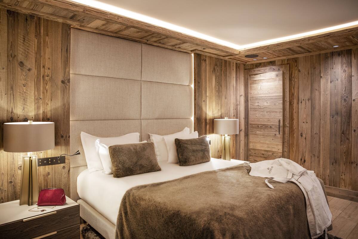 Habitación de hotel con pared de paneles de madera, una cama grande con ropa blanca, dos lámparas de noche y un bolso rojo sobre la mesita.