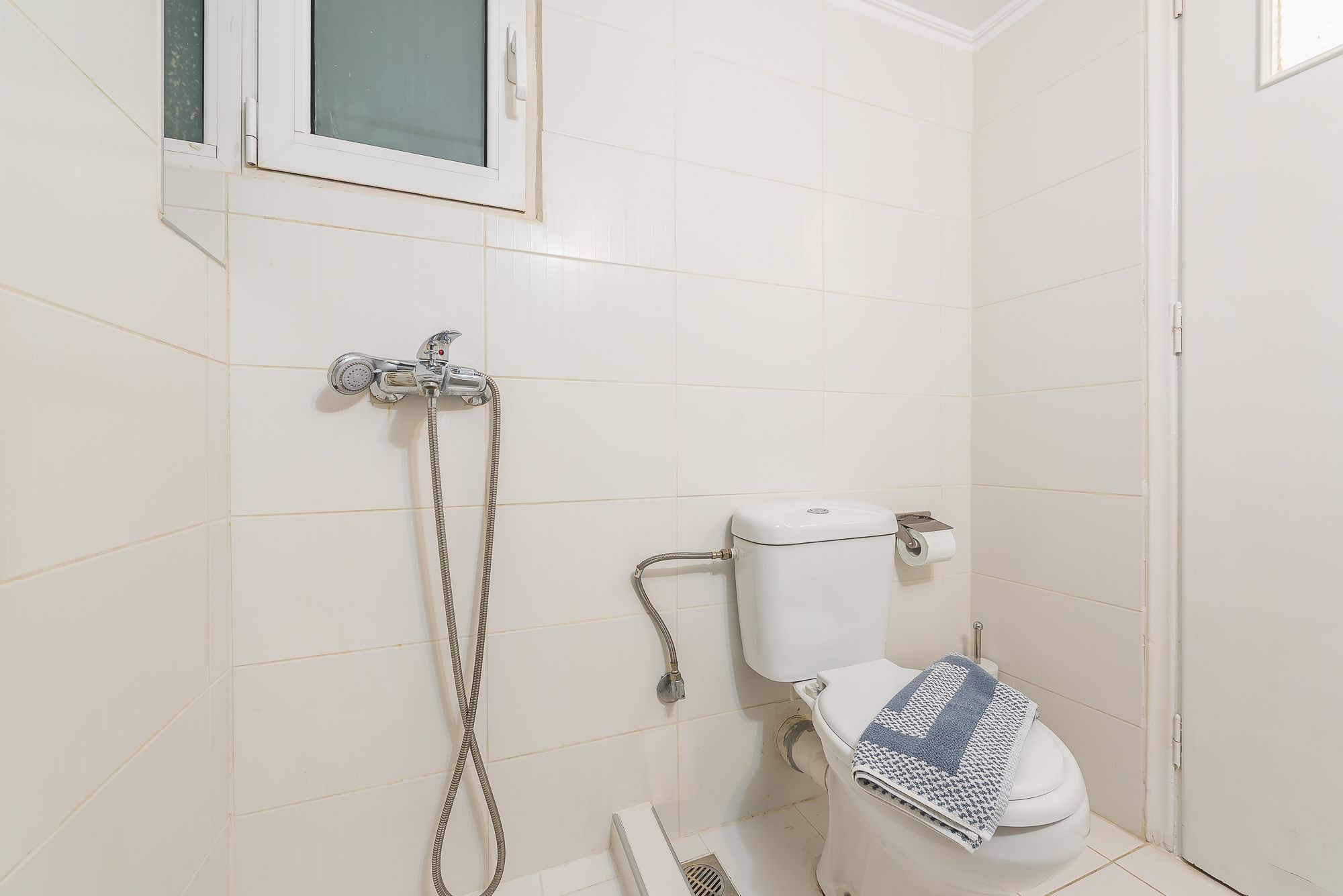 A small, clean bathroom with white tiles, a toilet, a showerhead, and a window.