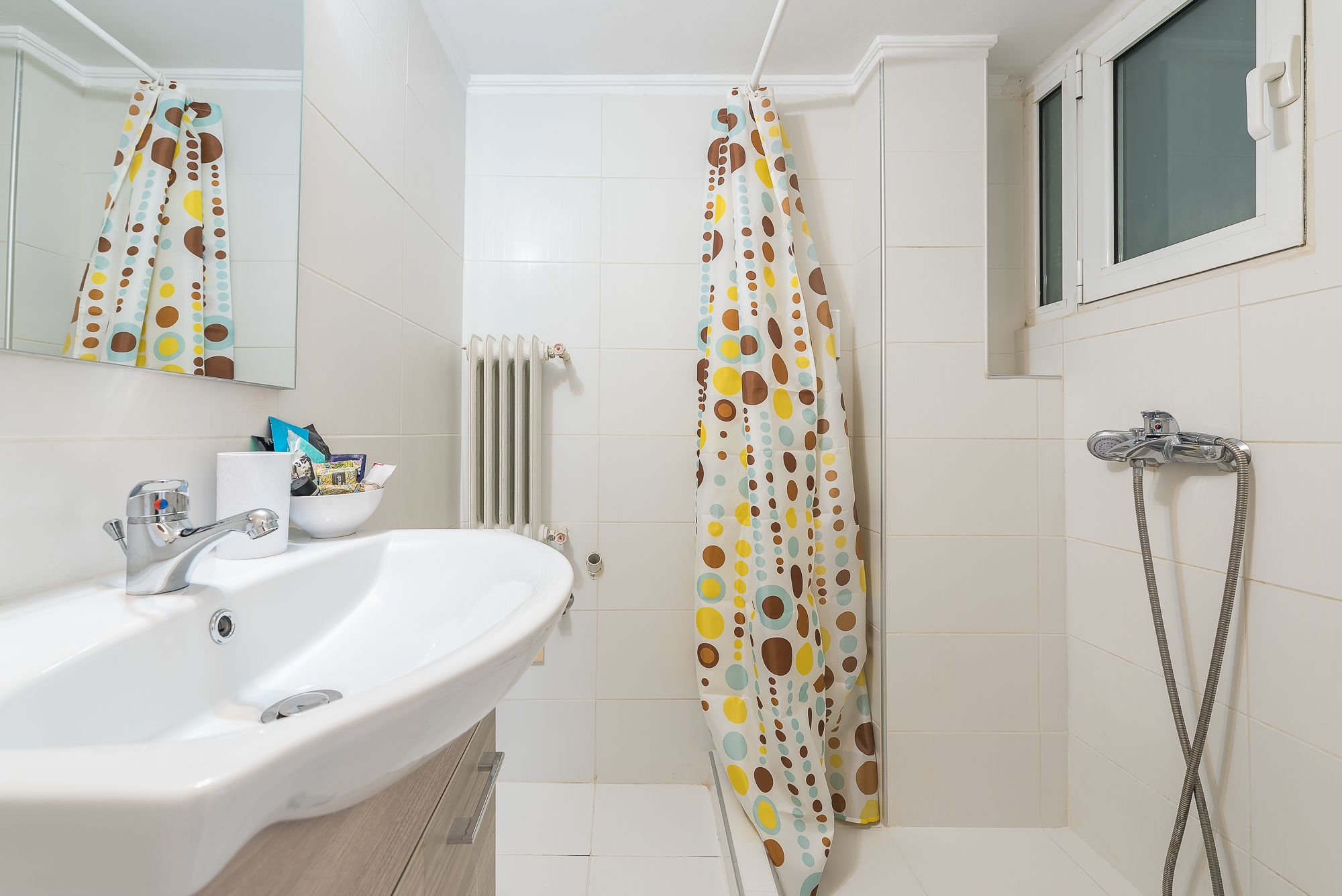 A bathroom with a white sink, a mirror, a shower curtain with a polka dot pattern, a showerhead, and a window.