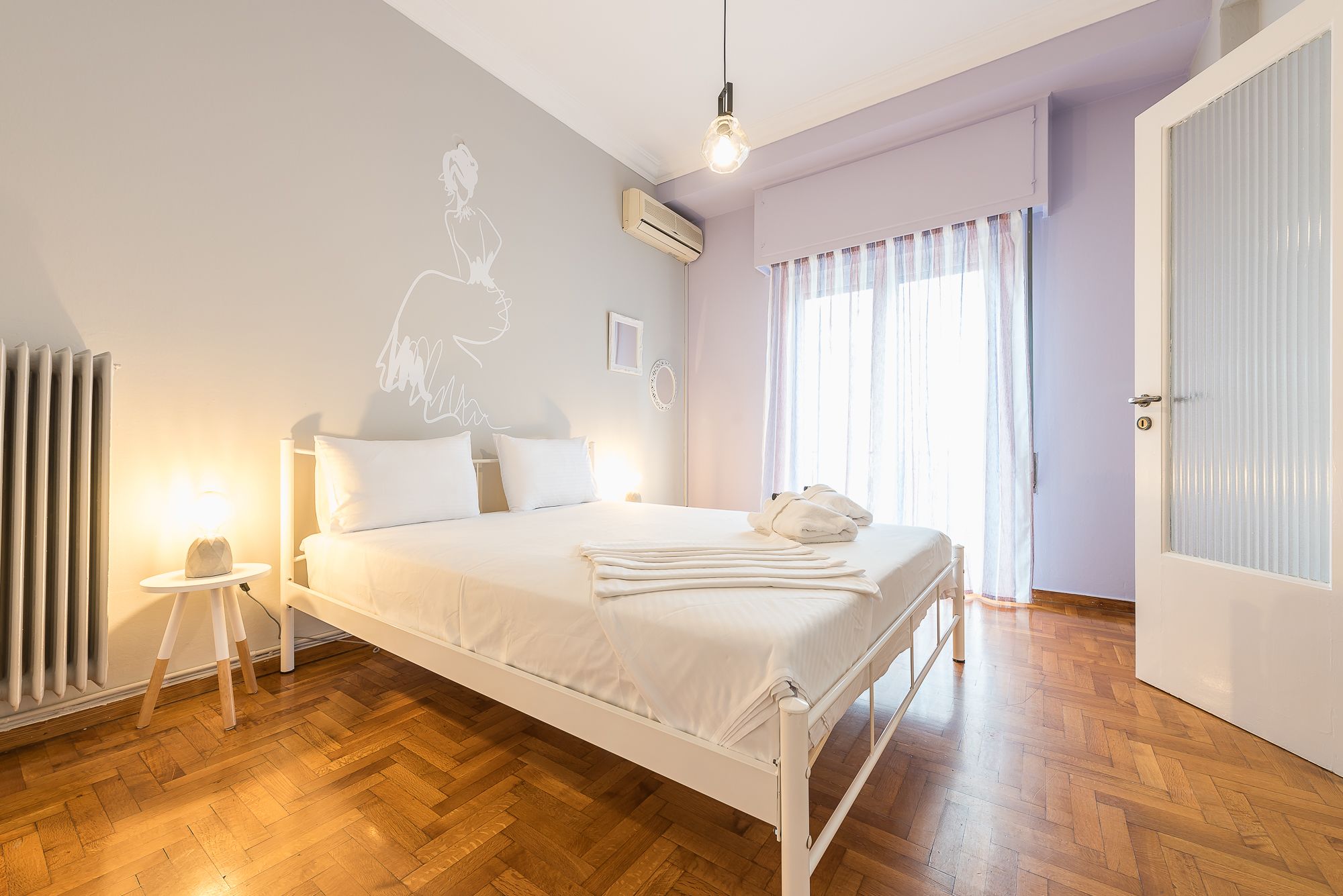 A bedroom with a bed, bedside tables, a radiator, an air conditioning unit, a window with curtains, and a door.