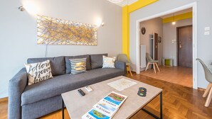 Ein modernes Wohnzimmer mit einem grauen Sofa, einem Couchtisch mit Büchern und einer Fernbedienung, einem Wandbild und einem Flur, der in ein weiteres Zimmer führt.