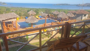 Un espace piscine agrémenté de parasols en chaume et de chaises longues en bois, offrant une vue sur une étendue d’eau et une végétation luxuriante.