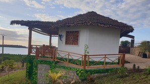 Une petite construction à un seul étage, dotée d’un toit de chaume, d’une terrasse en bois et offrant une vue sur une étendue d’eau en arrière-plan.