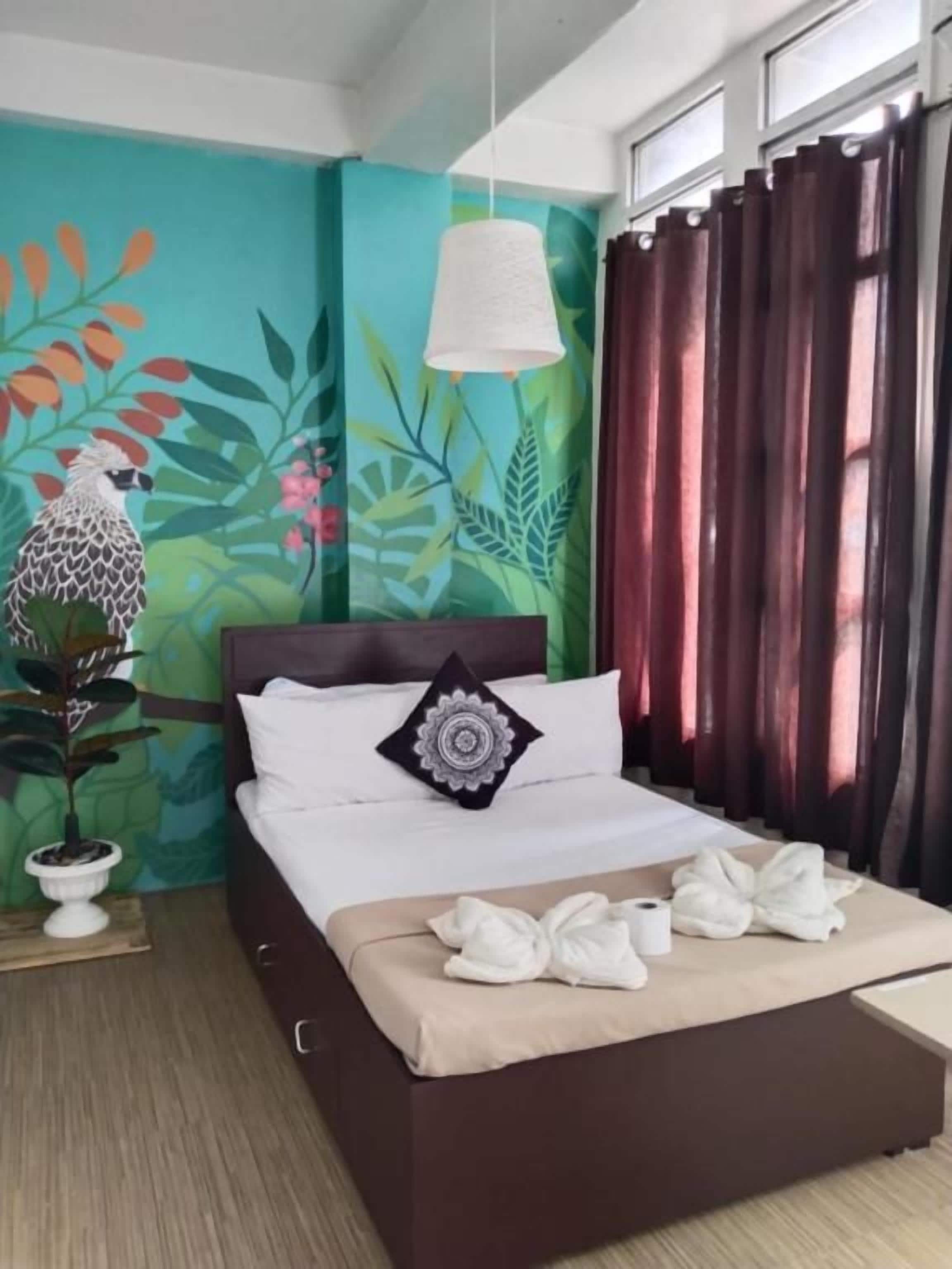 A hotel room with a bed, a potted plant, a wall mural, and a window with curtains.