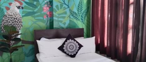 A hotel room with a bed, a potted plant, a wall mural, and a window with curtains.