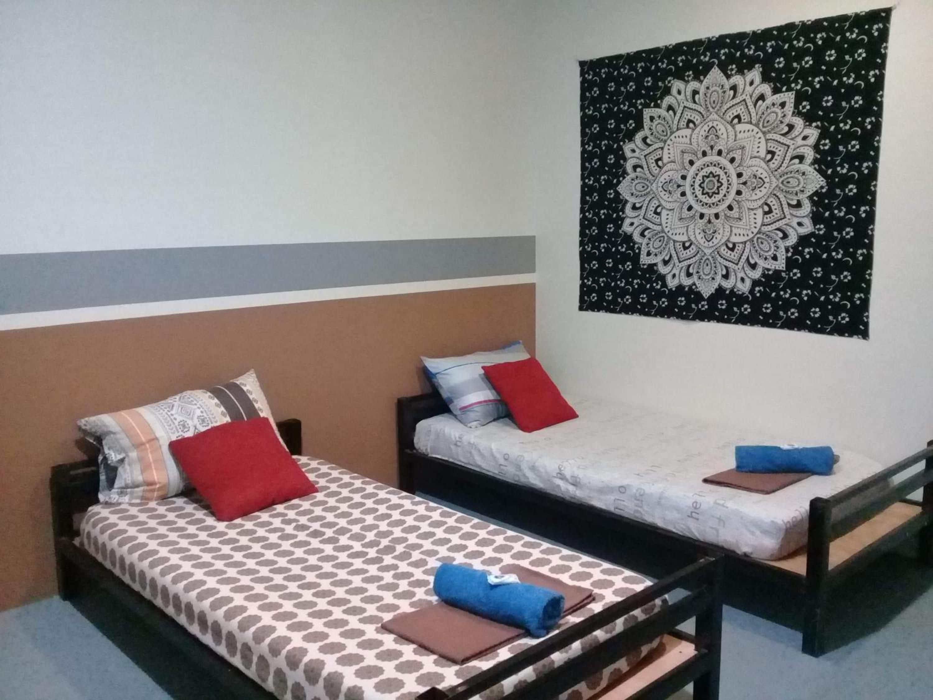 A room with two single beds, each with a patterned bedspread and pillows, a wall tapestry, and a plain wall.