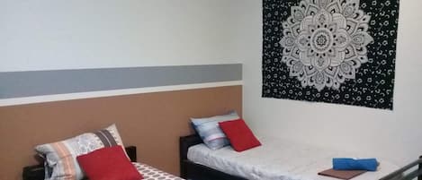 A room with two single beds, each with a patterned bedspread and pillows, a wall tapestry, and a plain wall.