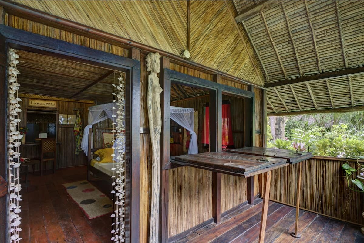 A rustic wooden room with a bed, a small table, and a view of greenery outside.