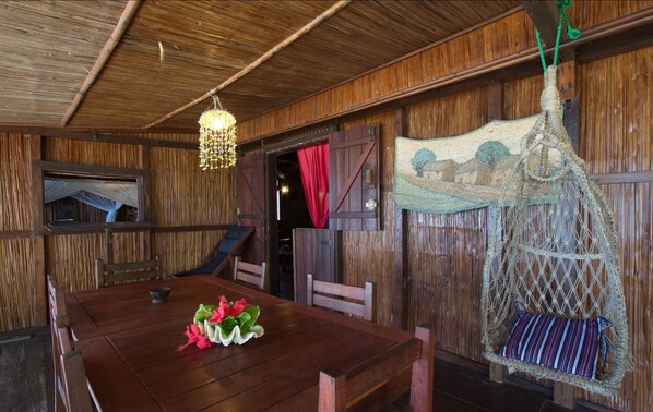 A rustic wooden room with a dining table, chairs, a hanging light fixture, and a hammock. - Analatsara Eco Lodge (Nosy Nato)
